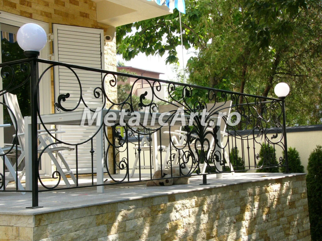 Balustradă exterioară pe zid de piatră, cu volute repetate și stâlpi cu glob de iluminat.