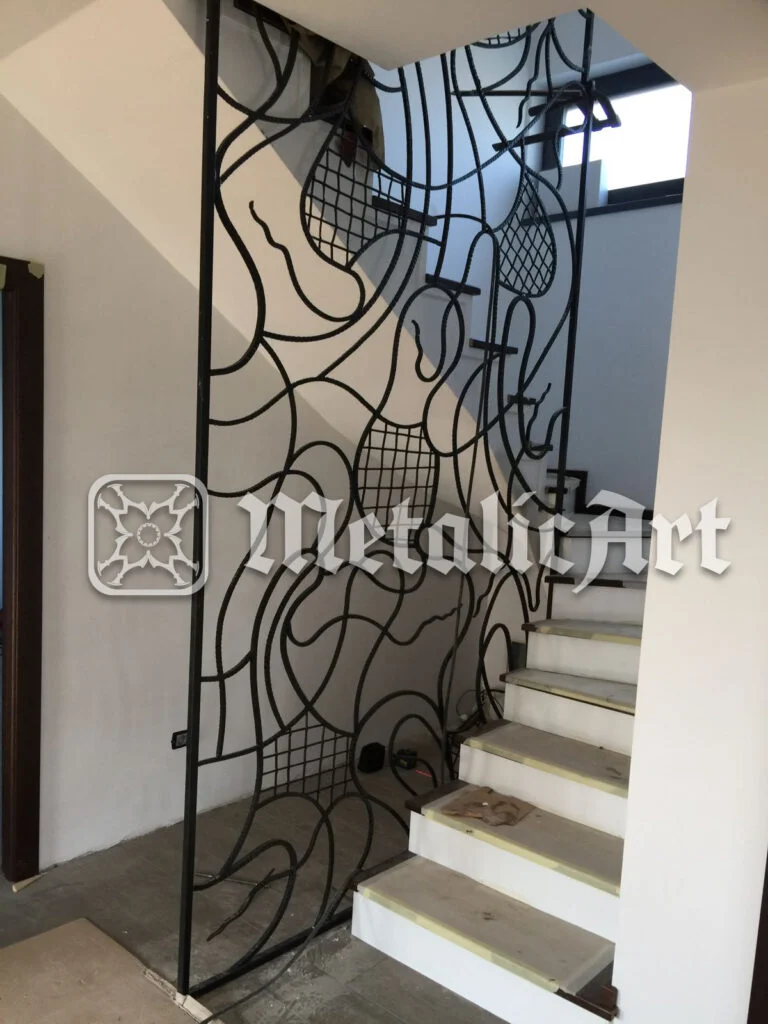 Panou-balustradă înalt, cu linii curbe și zone de plasă, la intrarea către scară.