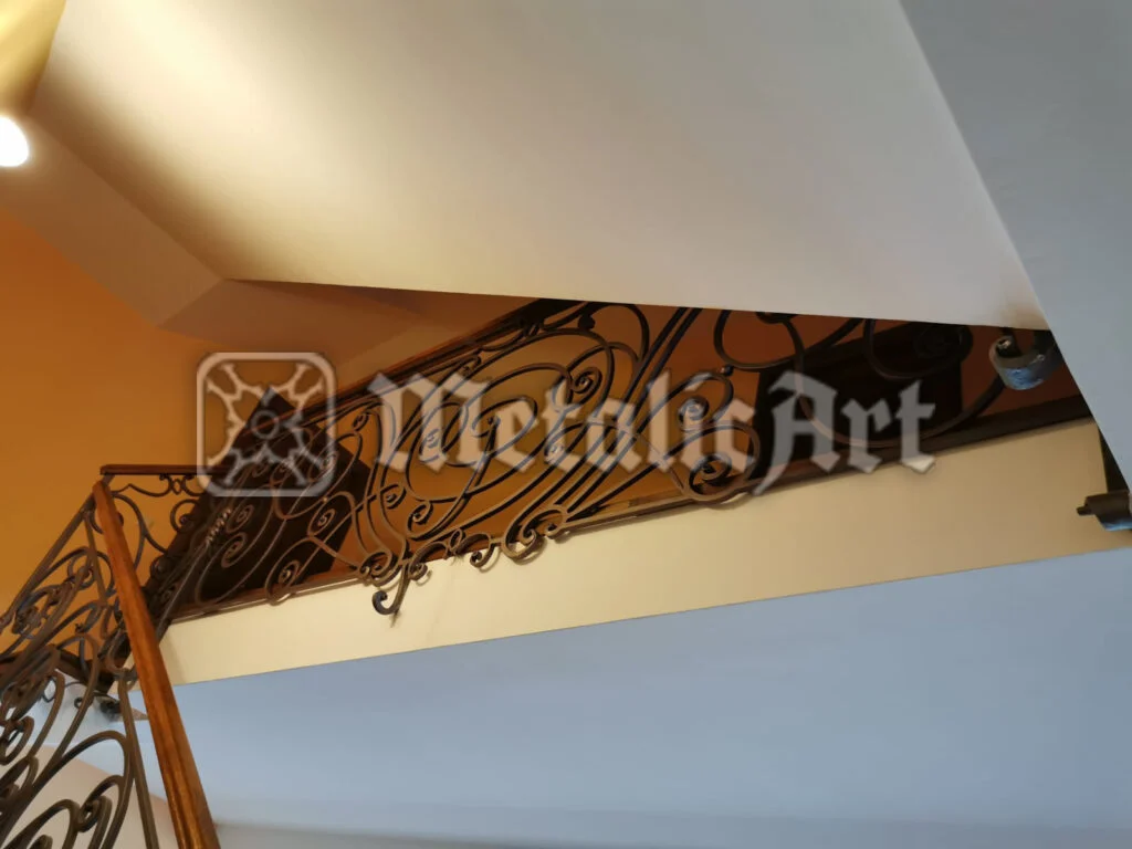 Balustradă de scară cu desen dens de volute, văzută de sub rampă, cu mână curentă din lemn.