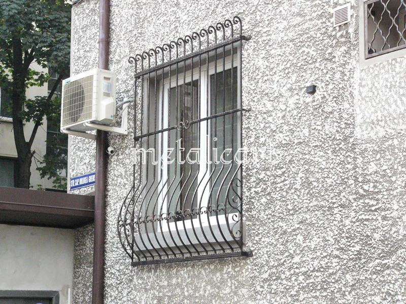 Grilaj bombat cu bare ondulate și mici volute pe margini, pe o fațadă cu tencuială aspră.