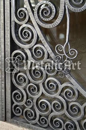 Detaliu de grilaj cu spirale dense, în finisaj argintiu-lucios, pe fundal de geam.
