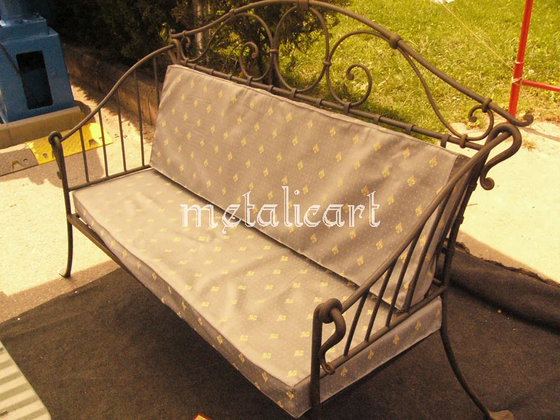 Canapea forjată cu spătar din volute și saltea deschisă la culoare, în curte.