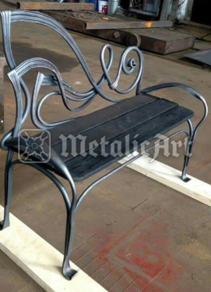 Bancă forjată cu structură din bare curbate și șezut din scânduri, în atelier.
