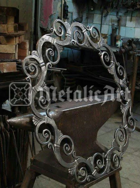 Ramă de oglindă forjată, cu volute și frunze, fotografiată pe nicovală în atelier.