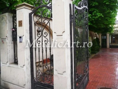 Poartă pietonală cu volute și bare răsucite, între stâlpi de beton profilat.
