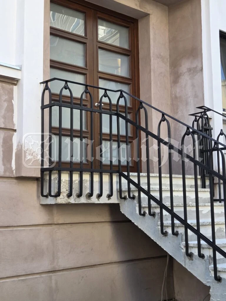 Rampă de scară din fier forjat cu arcade ogivale, de-a lungul fațadei, sub o ușă de lemn.