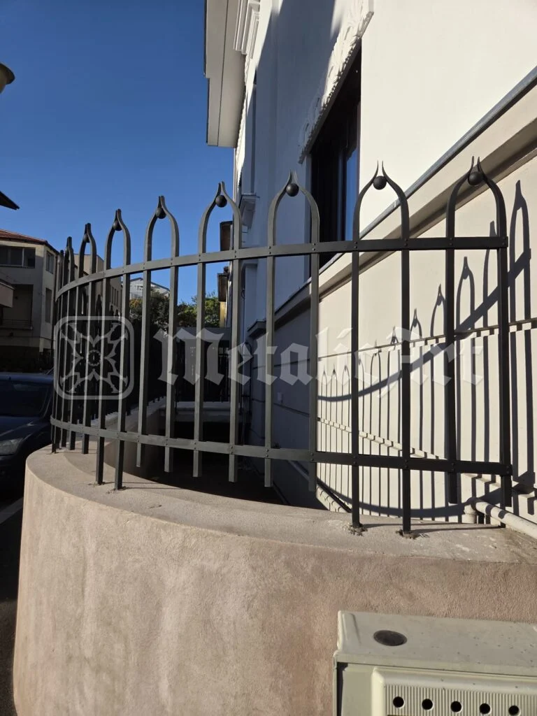 Balustradă curbată cu arcade ogivale și muguri forjați, în lumina soarelui.