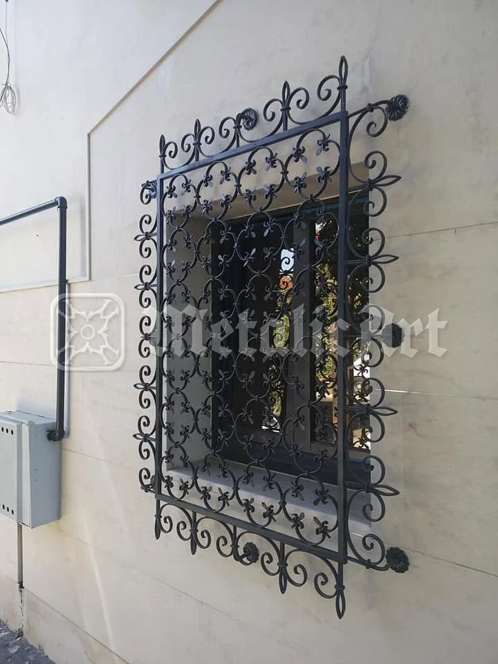Grilaj cu împletitură deasă și ramă de volute, la fereastra casei placate cu piatră.