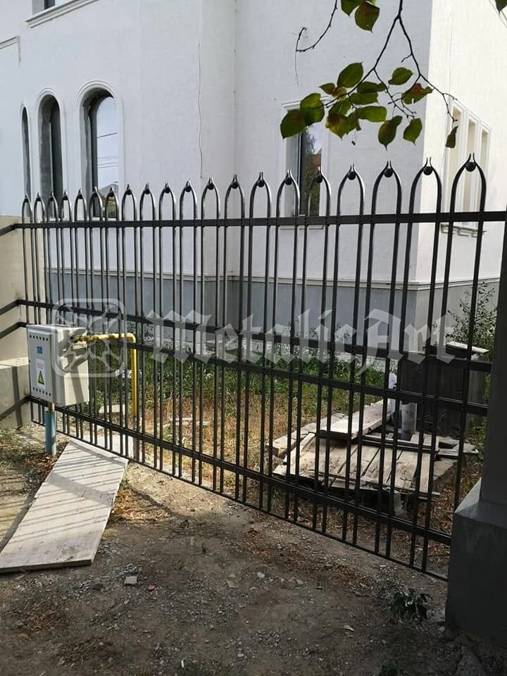 Gardul cu bare cu vârfuri ogivale, în fața casei cu ferestre arcuite.